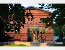 Backsteinfassade eines rechteckigen Gebäudes mit symmetrischen Mustern und kleinen Fenstern, davor ein großer Olivenbaum in einem Gefäß; umgeben von grünem Rasen und Schatten von Bäumen.