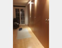 Verschwommenes Bild eines Wohnraums mit Holzboden, großen Einbauschränken auf der rechten Seite und einer Glastür mit Rahmen im Hintergrund. Ein Teppich und Hausschuhe sind auf dem Boden zu sehen.