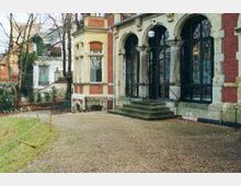 Historisches Gebäude mit roten Backsteinwänden und dekorativen Rundbögen über großen schwarzen Fenstern und Türen; eine kleine Treppe führt vom Kiesweg zum Eingang, umgeben von einer Grünfläche und Bäumen im Hintergrund.