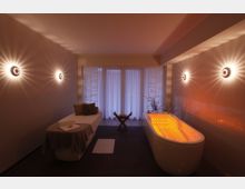 Wellnessraum mit einer beleuchteten Whirlpool-Badewanne und einer Liege. Der Raum ist sanft beleuchtet mit Wandleuchten, die ein strahlenförmiges Lichtmuster erzeugen, und einem großen Fenster mit halbtransparenten Vorhängen im Hintergrund.