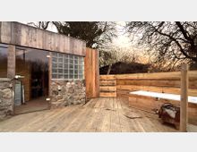 Holzterrasse im Außenbereich neben einem kleinen Gebäude mit Holzfassade, Steinsockel, Glastür und einem Fenster aus Glasbausteinen. Rechts steht eine lange, weiß verkleidete Bank vor einem hohen Holzsichtschutz; im Hintergrund sind kahle Bäume zu sehen.