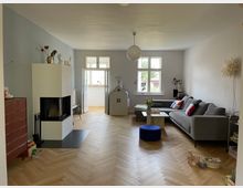 Wohnzimmer mit hellem Fischgrätenparkett, grau-blauer Wand, einem modernen Kamin links und einer großen grauen Eckcouch rechts. Ein Spielhaus und Spielsachen sowie ein Beistelltisch mit Blumen befinden sich im Raum, der von Tageslicht durch zwei Fenster und eine geöffnete Balkontür beleuchtet wird.