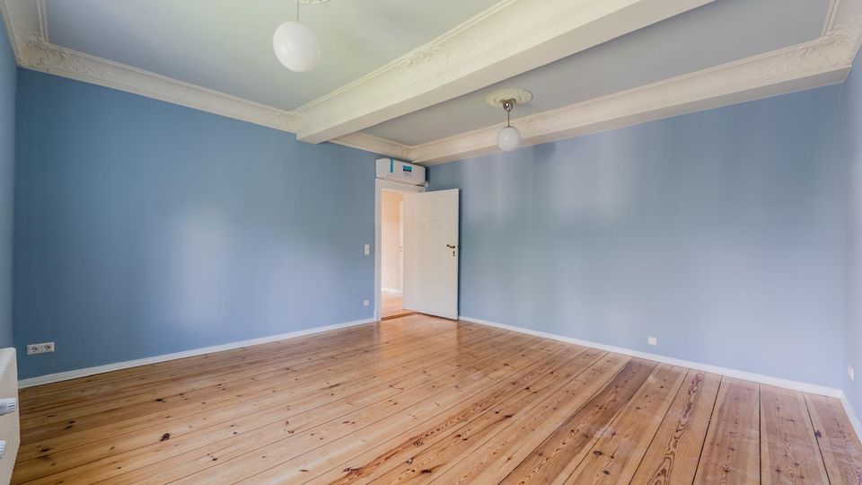 Leerer Innenraum mit hellblauen Wänden und breiten, lackierten Holzdielen. Weiße Stuckdecken mit Querbalken und zwei runden Deckenleuchten sowie eine offene Tür zu einem angrenzenden Flur.