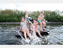 Kinder spielen und spritzen Wasser auf einem hölzernen Steg in einem natürlichen Gewässer, umgeben von Schilf und Pflanzen. Einige Kinder tragen Badeanzüge, während sie auf dem Steg stehen oder sitzen.