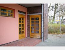 Eingangsbereich eines Gebäudes mit zwei Holztüren und einem kleinen Fenster. Die Fassade ist teils rosa verputzt und teils aus Backstein, umgeben von einem gepflasterten Vorplatz und kahlen Bäumen im Hintergrund.