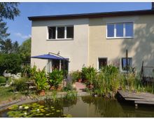 Zweistöckiges Wohnhaus mit heller Fassade, großen Fenstern und einem Garten. Im Vordergrund befindet sich ein Teich mit Wasserpflanzen und Seerosen, daneben stehen Terrassenmöbel, ein Sonnenschirm und Blumentöpfe.