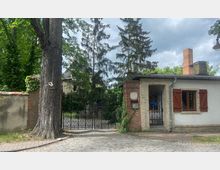 Eine gepflasterte Einfahrt mit einem schwarzen, verzierten Eisentor, flankiert von gemauerten Pfeilern und einer angrenzenden Hausfassade in weißer und roter Backsteinoptik. Im Hintergrund sind Bäume und ein großes Gebäude zu sehen, und ein Baum mit grober Rinde steht links vor einer niedrigen, überwucherten Mauer.