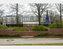 Öffentliche Toilettenanlage aus roten Ziegelsteinen, umgeben von niedrigen Hecken und Gehwegen, mit Beschilderung für "Herren" und "Damen"; im Hintergrund stehen Bäume und moderne mehrstöckige Gebäude.