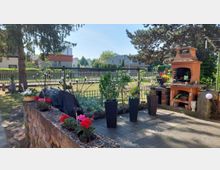 Gepflasterte Gartenterrasse mit gemauertem Außenkamin und Grill, mehreren hohen Pflanzkübeln und einem niedrigen Steinbeet mit Blumen. Im Hintergrund stehen Rasenfläche, Zaun, Bäume und Wohnhäuser unter blauem Himmel.