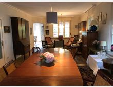 Esszimmer mit einem großen, rechteckigen Holztisch und mehreren Stühlen, umgeben von antiken Möbeln wie einem Schrank und einer Kommode. Der Raum ist mit Teppichen, Lampen und Dekorationen wie Bildern an den Wänden gestaltet und hat große Fenster, die Tageslicht einfallen lassen.