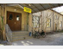 Eingangsbereich eines einfachen Gebäudes mit Holz-Doppeltür und einem gelben Schild mit der Aufschrift "Theater Café", darüber ein provisorisches Metalldach. Im Vordergrund sind zwei Fahrräder an ein Geländer gelehnt, und die Umgebung zeigt kaum bepflanzte Erde und Pflastersteine.