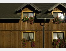 Holzhausfassade mit dunklem Schindeldach, mehreren Fenstern mit weißen Gardinen und Blumenkästen, die mit roten und pinken Geranien bepflanzt sind. Regenrinnen verlaufen entlang der Fassade.