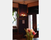 Innenraum eines gemütlichen Cafés mit dunkelvioletter Wand, einer antiken Wandleuchte und großen Fenstern mit Holzeinfassungen. Im Vordergrund ist ein Blumenstrauß mit Tulpen auf einem Tisch zu sehen.