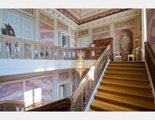 Prunkvoller Treppenhausbereich mit hölzernem Treppengeländer, verzierten Relieftafeln und dekorativen Malereien an den Wänden. Eine hängende, goldene Laterne und klassische Statuen unterstreichen die elegante Innenarchitektur.