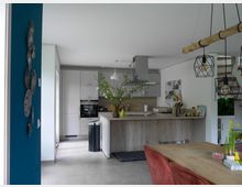 Moderne Wohnküche mit einer Kücheninsel aus Holz, weißer Einbauküche und Dunstabzugshaube. Im Vordergrund ein Essbereich mit Holztisch, roten Stühlen und Dekorationen, im Hintergrund ein Treppenaufgang und helle, minimalistische Gestaltung.