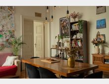 Cozy apartment dining-living room with a long wooden table and dark chairs under hanging pendant bulbs, opening to white paneled double doors. The space features a tall bookcase, framed art and drawings on the walls, potted plants, and a wooden sideboard with vases of dried flowers.