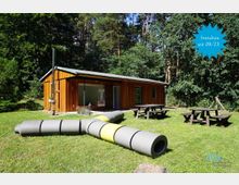 Ein eingeschossiges, modernes Holzgebäude inmitten einer Waldlandschaft, umgeben von Rasenflächen. Im Vordergrund befinden sich mehrere Picknicktische sowie große, röhrenförmige Spielgeräte auf der Wiese.