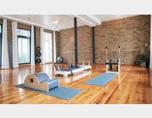 Heller Trainingsraum mit Holzboden, freigelegter Backsteinwand und großen Fenstern mit weißen Vorhängen. Im Raum befinden sich Pilatesgeräte, Matten und Fitnesszubehör, umgeben von einer minimalistisch eingerichteten Umgebung.