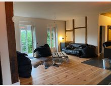 Wohnzimmer mit offenem, modern-rustikalem Design. Sichtbare Holzbalken an weiß verputzten Wänden, Holzboden, schwarze Sitzmöbel und ein Couchtisch aus einer Holzpalette; zwei Flügeltüren führen auf eine Terrasse mit Blick ins Grüne.