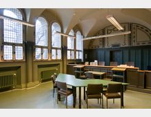 Innenraum eines historischen Arbeitszimmers mit hohen Rundbogenfenstern, Verglasung im Sprossenstil und dunkelgrünen Holzvertäfelungen. In der Mitte befinden sich grüne Tische und Stühle, während im Hintergrund ein Empfangstresen mit Dekorationselementen an der Wand sichtbar ist.