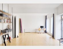 Heller, minimalistischer Arbeitsraum mit Holzboden, einem Regal an der linken Wand, bunten Stoffrollen in einer Ecke und einem Arbeitsplatz mit zwei Stühlen und einem Monitor vor großen Fenstern rechts.