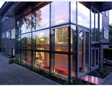 Modernes Gebäude mit einer Glasfassade, die einen Blick ins Innere ermöglicht, wo ein beleuchtetes Treppenhaus mit rotem Akzent sichtbar ist. Das Gebäude ist an ein älteres Backsteinhaus angebaut, umgeben von einem Kiesweg und Pflanzen.