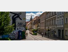 Straßenszene in einer Altstadt mit einer Straßenbahn auf Schienen, umgeben von historischen Gebäuden mit verzierten Fassaden. Auf einer Gebäudewand links ein großes Wandgemälde und Graffiti, im Hintergrund ein Kirchturm mit Uhr.
