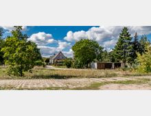 Wohnsiedlung mit Einfamilienhäusern und Gärten, umgeben von Bäumen und Wildwuchs; im Vordergrund eine gepflasterte Fläche mit Grasbewuchs und ein einfacher, flacher Garagenkomplex.