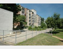 Mehrfamilienhaus-Siedlung mit vierstöckigen Gebäuden in schlichter Bauweise, teils aus Ziegeln und Beton, umgeben von Grünflächen mit Bäumen und Rasen. Im Vordergrund befindet sich ein Gehweg mit Metallgeländer und gepflastertem Zugang.