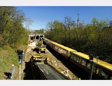 Baustelle an einer Zugstrecke mit Arbeiten an einem Tunnelausgang, mehreren Baufahrzeugen und Arbeitern. Rechts steht eine gelbe S-Bahn; die Umgebung ist von Bäumen und Vegetation geprägt.