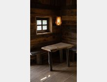 Rustikaler Innenraum mit Holzwänden, einer kleinen Fensternische mit Kreuzsprossen und einem hölzernen Tisch mit unregelmäßiger Oberfläche, flankiert von einfachen Holzbänken. Eine Hängelampe mit warmem Licht beleuchtet den Raum.