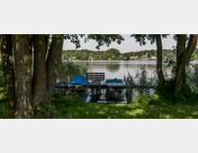 Ein ruhiger See mit einem schmalen Bootssteg, auf dem mehrere Boote mit Planen abgedeckt liegen, umgeben von Schilf und Bäumen im Vordergrund. Im Hintergrund sind bewaldete Ufer und vereinzelte Gebäude sichtbar.