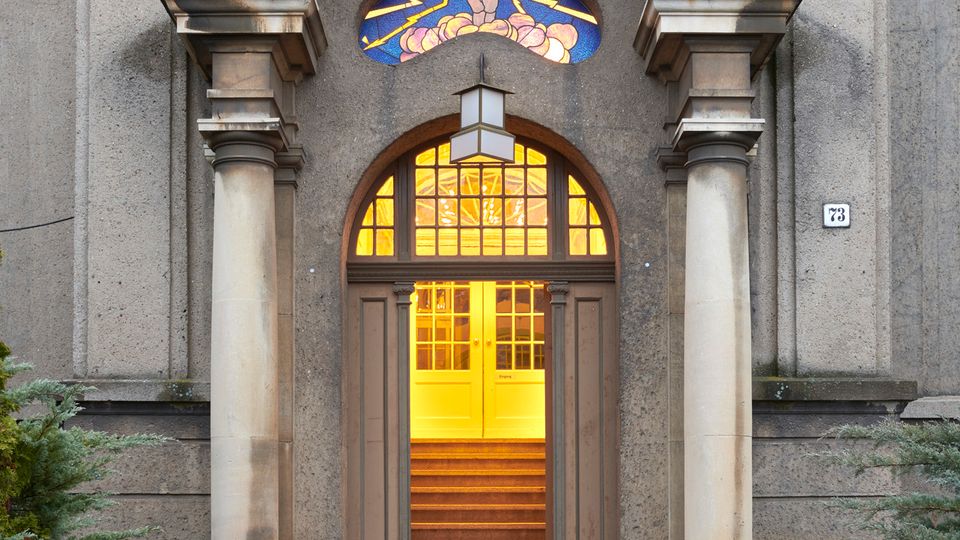 Eingang eines historischen Gebäudes mit zwei Säulen, einem gewölbten Türbogen und einem farbigen Bleiglasfenster mit religiösem Motiv. Die beleuchtete Tür führt zu einer Treppe, die ins Innere des Gebäudes führt, umgeben von einem gepflasterten Außenbereich mit Grünpflanzen.