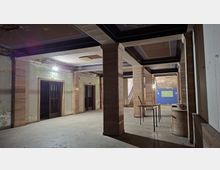 Große, unrenovierte Innenhalle eines älteren Gebäudes mit breiten Säulen, dunklen Deckenbalken und abblätterndem Putz an den Wänden. Links befinden sich doppelflügelige Türen, im Hintergrund eine Treppe, und rechts stehen provisorische Tische sowie Baumaterialien.