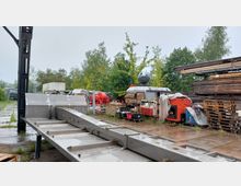 Ein regnerischer Außenbereich mit verschiedenen Maschinen, Holzstapeln und Metallstrukturen. Im Hintergrund stehen Bäume und Container, während der Boden aus Betonplatten besteht.