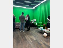 Indoor studio space with green-screen fabric covering the walls and bright fluorescent strip lights and acoustic foam panels on the ceiling. The room has wood-look flooring, a few chairs and tables with party decorations, balloons, and packaged supplies.