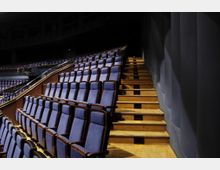 Innenraum eines Theaters oder Auditoriums mit mehreren Reihen gepolsterter Stühle in Dunkelblau, angeordnet auf ansteigenden Holztribünen. Rechts im Bild befindet sich eine Treppe aus Holz, die entlang einer dunklen Wand mit Säulen verläuft.