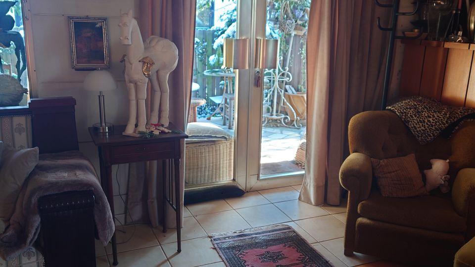 Wohnzimmer mit gemütlichem Ambiente, einem Mix aus Holzmöbeln und Teppichen in warmen Farben. Große Glastüren führen zu einer Terrasse mit Pflanzen; der Raum ist durch Gemälde, Vorhänge und dekorative Elemente gestaltet.