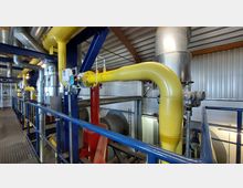 Innenansicht einer Industrieanlage mit gelben und silbernen Rohren, blauen Stützsäulen und einer Metallplattform. Im Hintergrund sind Lüftungsschächte, Metallwände und weitere Rohrleitungen sichtbar.