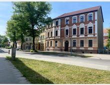 Straßenszene mit einem dreigeschossigen Wohngebäude im historischen Stil, das durch rote Ziegel und weiße Fassadenelemente geprägt ist. Im Vordergrund eine Grünfläche, flankiert von Gehweg und Straße, mit mehreren Bäumen entlang der Häuserreihe.