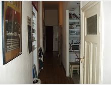 Schmaler Flur in einer Wohnung mit Holzboden, weiß gestrichenen Wänden und einer geöffneten Tür im Vordergrund. Links befinden sich ein Wandposter und Regale, rechts ein Einbauschrank mit Büchern und Geschirr.