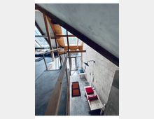 Blick in einen hohen, loftartigen Innenraum mit sichtbaren Holzbalken, geneigter Decke und großem Dachfenster sowie bodentiefen Glasfronten am Ende. Eine schmale Galerie mit Holzgeländer führt entlang der Wand; unten stehen wenige Möbel auf Betonboden vor einer hellen Ziegelwand.