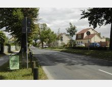 Straßenszene in einem Wohngebiet mit Einfamilienhäusern, Vorgärten und einer schmalen Fahrbahn, gesäumt von Bäumen und Verkehrszeichen, darunter Geschwindigkeitsbegrenzungsschilder für 30 und 50 km/h. Im Vordergrund ein Gehweg mit einem Plakatständer.