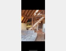 Wohnraum in einem rustikalen Dachgeschoss mit Holzbalken und schräger Decke. Im Vordergrund ein graues Ecksofa, eine Holztreppe führt zu einer offenen Galerie, und an der Wand ist ein Gemälde mit Landschaftsmotiv zu sehen.