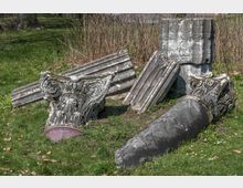 Fragmentierte antike Säulenelemente, darunter Kapitelle mit floralen Verzierungen und gebrochene Säulenteile, auf einer Wiese in einem parkähnlichen Umfeld. Im Hintergrund sind Sträucher und Bäume zu sehen.