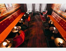 Schmaler Bar- oder Loungebereich mit langen, gepolsterten Lederbänken entlang beider Seiten, kleinen runden schwarzen Tischen mit Tischlampen und gemischten Polsterstühlen. Freiliegende Backsteinwände mit Bildern, dunkler Holzboden und bodenlange Vorhänge am Ende des Raums.