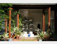 Offene Terrassentür eines Hauses mit Rahmen aus Holz, dahinter ein Regal mit Keramikgeschirr und dekorativen Kannen. Im Vordergrund stehen Topfpflanzen, darunter mehrere Sukkulenten, die den Eingangsbereich umrahmen.