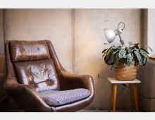 Gemütliche Sitzecke in einem Innenraum mit Sichtbetonwand: ein brauner Ledersessel mit gemustertem Sitzkissen steht neben einem kleinen Beistelltisch mit Korb-Topfpflanze und Schreibtischlampe. Rechts ist eine Fensterbank mit einfallendem Tageslicht zu sehen.