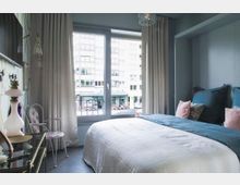 Ein kleines Schlafzimmer mit einem großen Fenster, das den Blick auf ein Gebäude und ein Straßencafé bietet. Das Zimmer ist mit einem Doppelbett, dekorativen Kissen, Vorhängen, einem Nachttisch sowie einem Stuhl mit floralem Muster ausgestattet.