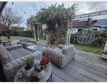 Terrasse mit grauen Rattanmöbeln, darunter Sessel und ein Tisch mit Glasplatte. Im Hintergrund befindet sich eine Pergola mit Kletterpflanzen, daneben ein hängender Rattansessel. Der Garten ist mit Rasen, Hecken und Bäumen gestaltet, während im Hintergrund Häuser und ein bewölkter Himmel zu sehen sind.